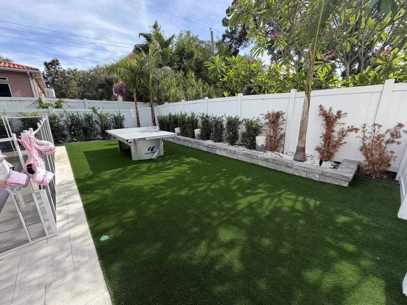 Family backyard of artificial turf with a ping-pong table along a white fence and tropical plantings