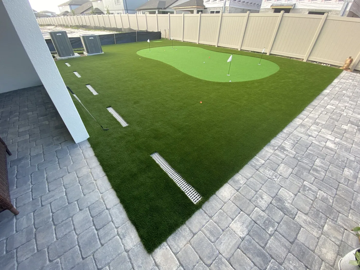 Backyard putting green with flags and a turf fringe, framed by gray paver walkways